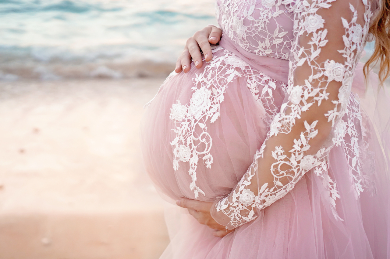 pregnant belly in pink lace dress with hands of mother on the background of sea. Coastal maternity photoshoot on the beach.